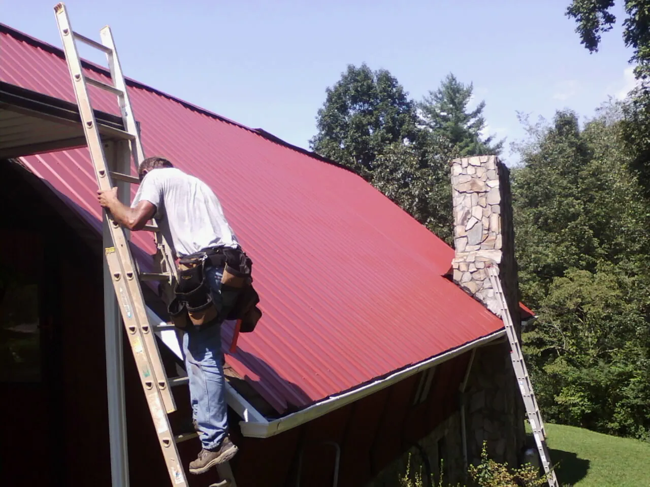 Roofer on a ladder beside a red metal roof and stone chimney — Soffit & Fascia Repair by Roofing Brothers in Beach City